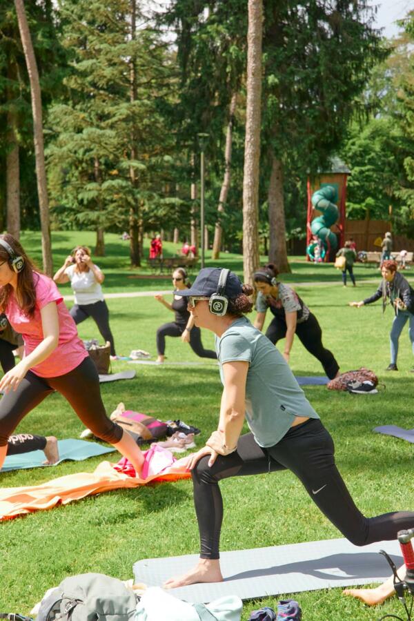 Eine Gruppe von Menschen nimmt an einer Yoga-Stunde im Freien teil und trägt Kopfhörer. Die Landschaft ist grün und von Bäumen umgeben.