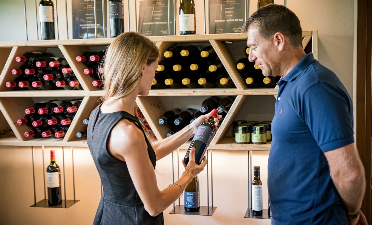 A woman shows a bottle of wine to a man in a shop. In the background, a shelf with various bottles of wine.