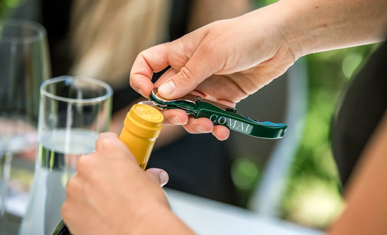 A hand using a corkscrew to open a bottle of wine. In the background, glasses and a convivial atmosphere can be seen.