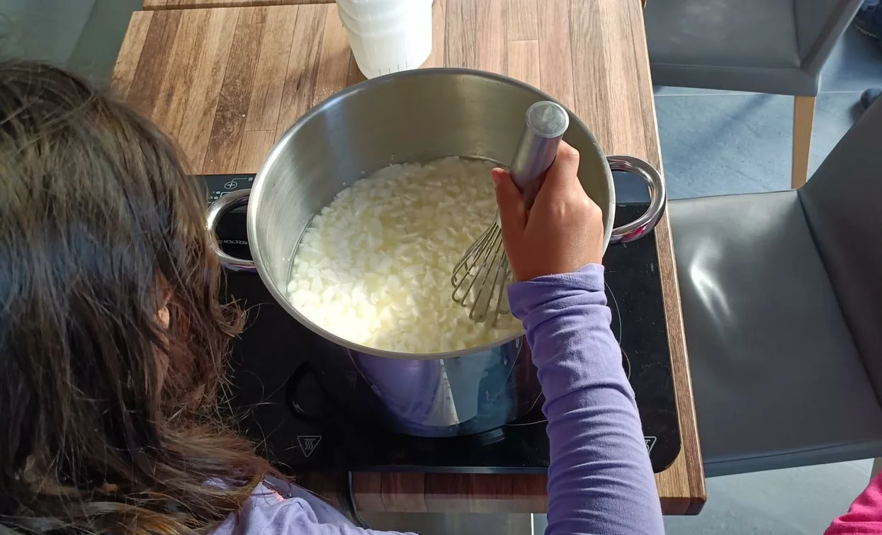 Una bambina mescola il composto in una pentola, mentre si trova a un tavolo. Sullo sfondo si vedono altre persone e dei bicchieri bianchi.