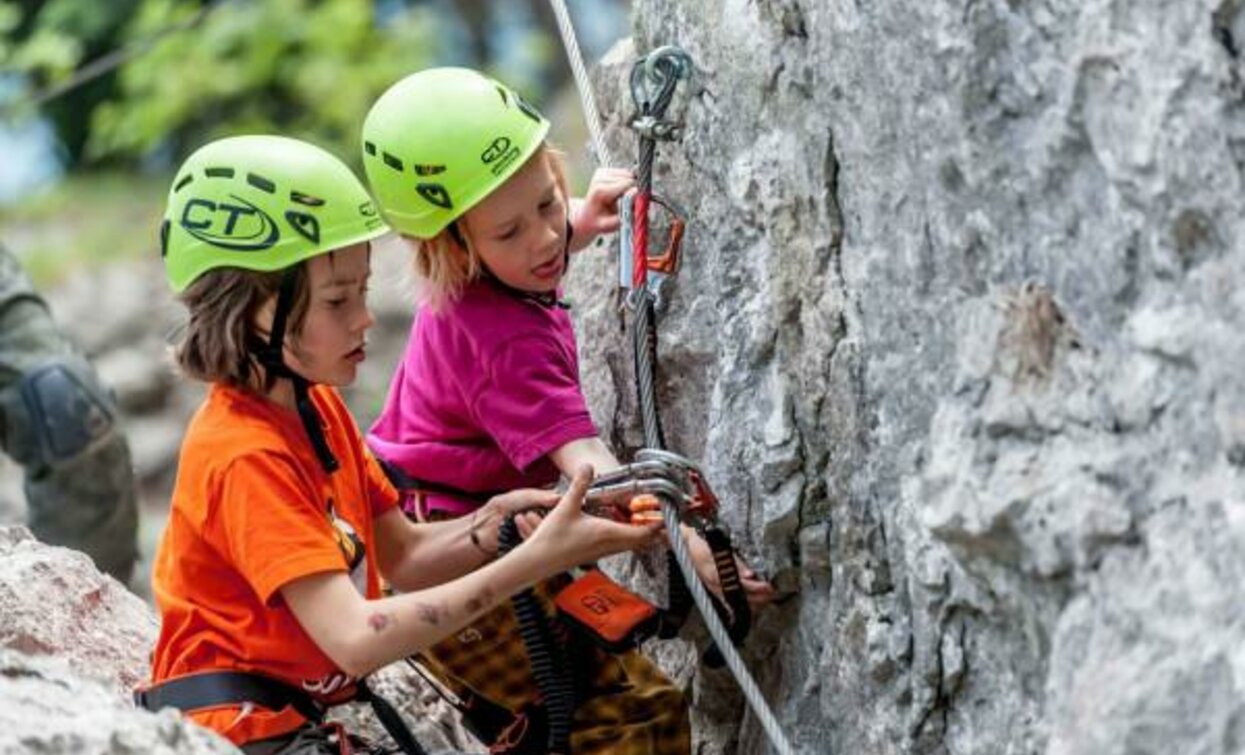 Zwei Jungs, die mit grünen Schutzkleidungen und Helmen gekleidet sind, klettern an einer Felswand. Einer von ihnen hilft dem anderen, die Sicherheitsausrüstung einzustellen.