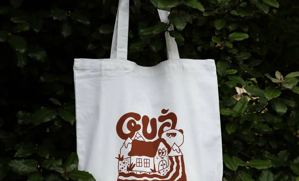 Una borsa di tela bianca appesa a un albero, con un design semplice e accattivante. La scritta "Guà" e l'immagine di una fattoria decorano il bag.
