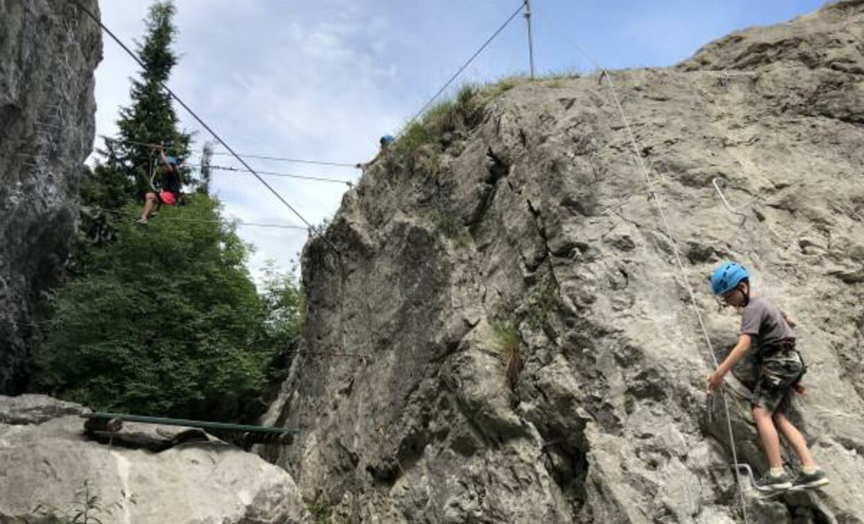 Un bambino scala una parete rocciosa, indossando un casco di sicurezza. Sullo sfondo, un adulto attraversa una corda sospesa tra le rocce.
