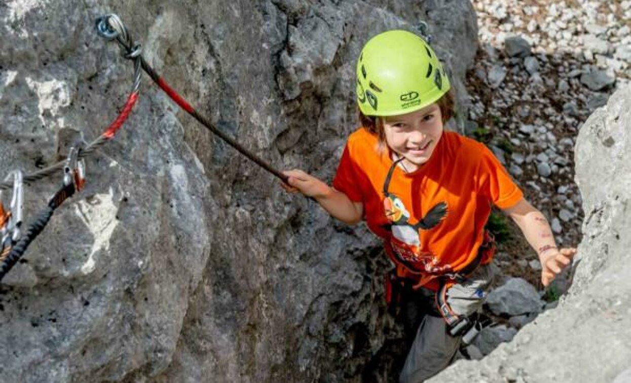 Un bambino sorridente sta arrampicandosi su una roccia, indossando un casco giallo. La scena mostra un ambiente roccioso, ideale per l'arrampicata.